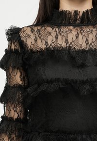 Femeie care poartă un top din dantelă neagră cu benzi orizontale cu volane și un guler înalt din dantelă, arătând mâneci din dantelă transparente și partea superioară a pieptului.