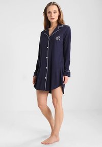 Kobieta w granatowej koszuli nocnej na guziki z białym wykończeniem, stojąca boso na jednolitym jasnym tle.