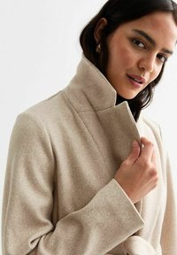 Abrigo de lana beige con un cuello alto y diseño envolvente, que presenta un lazo visible en la cintura y un acabado suave y texturizado.