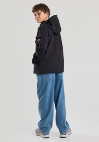 Black hooded jacket with zippers, side pockets, and a logo patch on the sleeve; worn with loose blue denim jeans and sneakers.