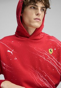 Sweat-shirt à capuche rouge avec un motif d'éclaboussures blanches, featuring un petit logo Puma et un badge Ferrari sur la poitrine. Matériau en mélange de coton.
