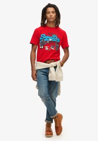 Rød bomulds t-shirt med grafisk print, lys blå tekst, lyseblå jeans og brune læderstøvler. Lys sweater bundet om taljen.