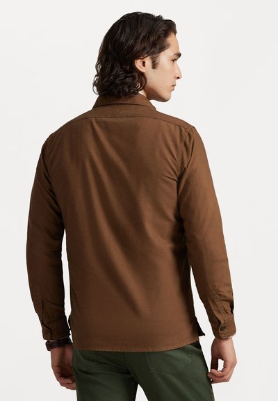 Camisa de manga larga marrón hecha de tela texturada, con un cuello clásico, puños con botón y un dobladillo recto. Detalles de costura visibles.