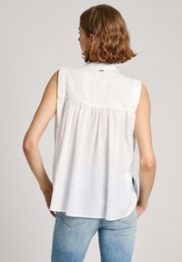 Hvid ærmeløs bluse med plisseret ryg, rund kant og en subtil hardware-detalje ved kraven. Kombinationen er med lyseblå jeans.