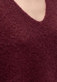 Gros plan d'un tissu doux et texturé de couleur rouge foncé d'un pull en V sur un fond de peau claire.
