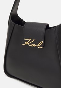 Sac en cuir noir avec un design structuré, arborant un accent en logo doré. Texture lisse avec un rabat magnétique plat.