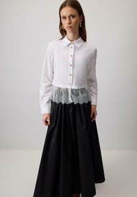 Bluză albă cu nasturi aurii și dantelă la talie, asociată cu o fustă lungă neagră cu pliseuri și o textură netedă.