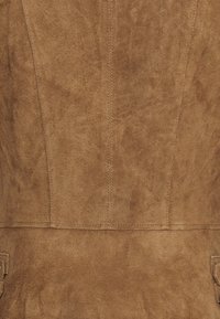Tessuto scamosciato marrone con una consistenza morbida, caratterizzato da cuciture verticali e piccole tasche laterali, creando una silhouette strutturata.