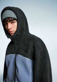 Jonge man met een grijze beanie en een zwart-blauwe fleece jack met ritssluiting en capuchon op, tegen een effen achtergrond.
