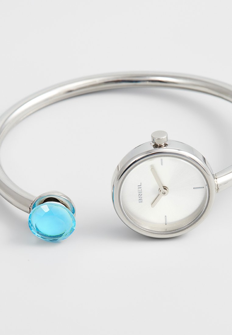 Zilveren bangle horloge met een witte wijzerplaat en minimale markeringen, met een blauwe ronde edelsteen aan de open uiteinde aan de andere zijde.