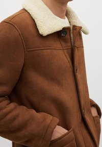 Brun ruskindsjakke med en shearling-krave, der har lynlåse foran, sidelommer og en glat overflade.
