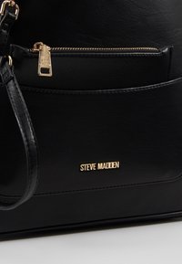 Sac à main en cuir noir avec des garnitures en métal doré. Comprend une poche zippée et le nom de la marque "Steve Madden" embossé à l'avant.