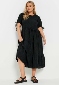 Svart, skiktad midiklänning med puffärmar och slipsdetaljer. Texturert tyg med en flödande siluett och ett sidoklyvning.