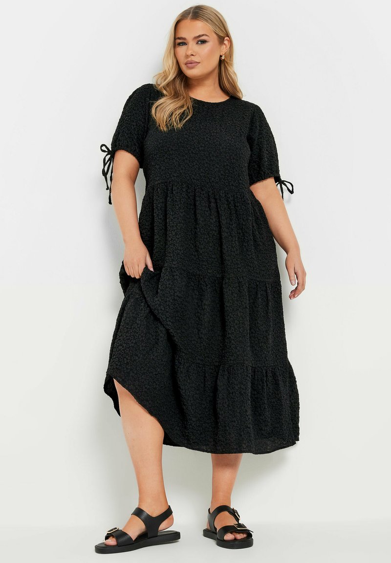 Svart, skiktad midiklänning med puffärmar och slipsdetaljer. Texturert tyg med en flödande siluett och ett sidoklyvning.