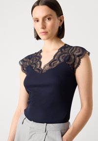 Débardeur marine à côtes avec des manches en dentelle noire transparente et un col en V. Texture lisse avec un motif de dentelle complexe le long du décolleté.