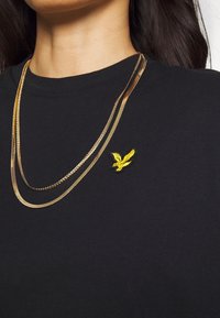 Zwarte shirt met een geborduurd embleem van een gouden adelaar, gelaagd met twee gouden kettingen van verschillende lengtes en texturen.