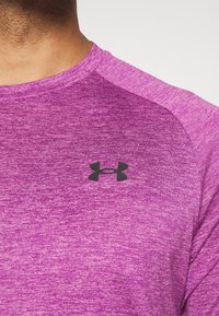 Detailní záběr osoby, která m á na sobě fialové sportovní tričko s černým logem Under Armour na hrudi.