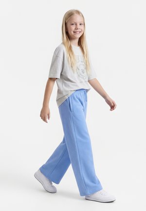 Jeune personne marchant, portant un t-shirt oversize gris clair, un pantalon ample bleu clair et des baskets blanches, souriante et regardant devant elle.