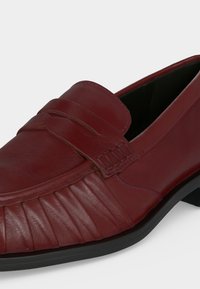 Ανδρικό σκούρο κόκκινο δερμάτινο loafer με λουράκι penny, πλισέ λεπτομέρεια μπροστά και μαύρη σόλα σε λευκό φόντο.