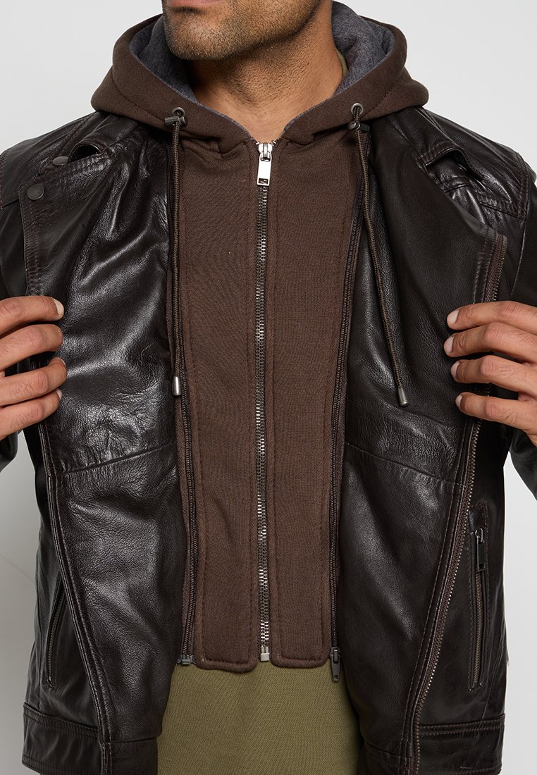 Chaqueta de cuero marrón con sudadera con cremallera en un tono marrón contrastante debajo. Cuenta con cierre de cremallera, capucha y dos bolsillos frontales. Textura suave.