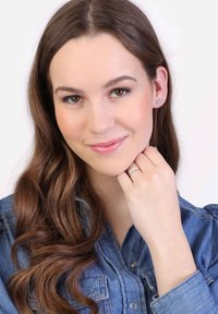 Vrouw met lang bruin haar die een denim overhemd draagt. Ze houdt haar hand dichtbij haar gezicht en toont een zilveren ring en stud oorbellen.