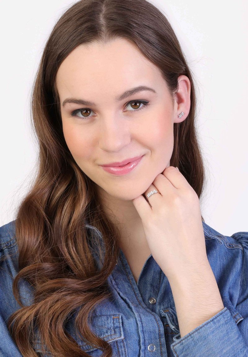 Vrouw met lang bruin haar die een denim overhemd draagt. Ze houdt haar hand dichtbij haar gezicht en toont een zilveren ring en stud oorbellen.