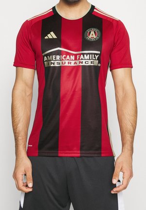 Mann trägt rotes und schwarzes Fußballtrikot von Atlanta United FC mit American Family Insurance-Logo und Adidas-Emblem, kombiniert mit schwarzen Shorts.