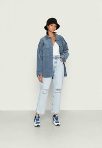 Camisa overshirt em veludo cotelê azul, top branco, jeans claros desfiados, chapéu bucket preto e ténis azuis com detalhes brancos. Roupa casual, pose em pé.