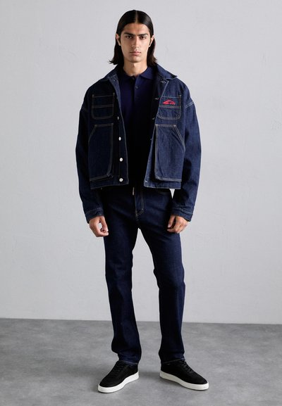 Denim jacket with two front pockets and contrasting yellow stitching, worn over a dark polo, paired with dark jeans and black sneakers.