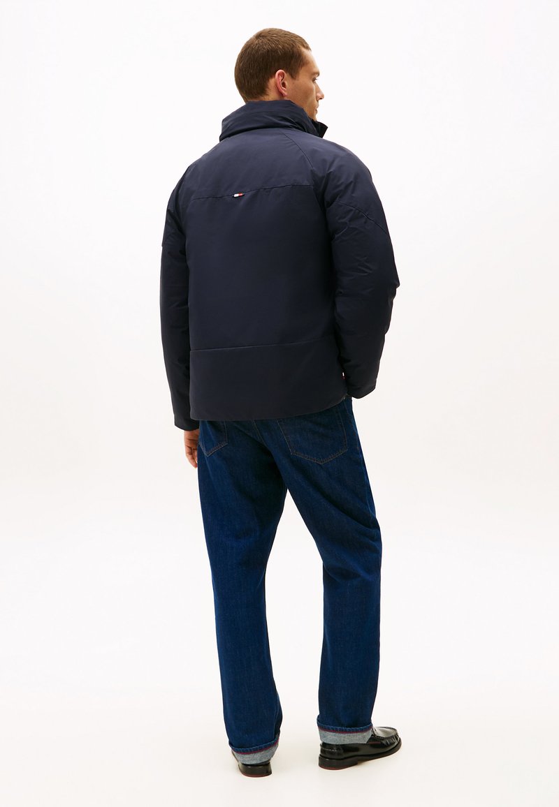 Mörkblå puffjacka med hög krage och liten logodetalj bak, kombinerad med mörkblå jeans och svarta skor.