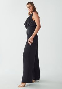 Combinaison noire avec un décolleté drapé, design sans manches et coupe pantalon évasée, fabriquée en tissu lisse et avec une taille cintrée.