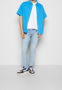 Camicia blu brillante a maniche corte con bottoni sopra una t-shirt bianca, abbinata a jeans slim-fit blu chiaro e sneaker nere con accenti blu.