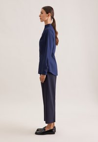 Navy blouse with a high collar, side buttons, paired with dark pants and black loafers, showcasing a streamlined silhouette and smooth fabric.