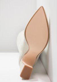 Escarpin beige à bout pointu avec semelle lisse et talon bloc, positionné à la verticale contre un mur et une surface blancs.