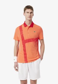 Polo arancione con design a strisce rosse e colletto rosso, con logo Lacoste verde. Indossato con pantaloni corti bianchi e un braccialetto.