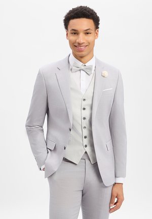 Jonge man die een lichtgrijs pak draagt, bijpassend vest en strikje, met een bloemenreversspeld, glimlachend tegen een effen witte achtergrond.