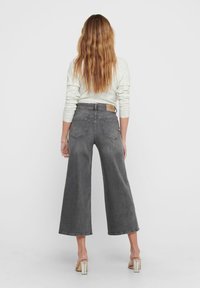 Nainen, jolla on pitkät laineikkaat hiukset, pukeutuneena valkoiseen neuletakkiin, korkeavyötäröisiin leveälahkeisiin harmaisiin farkkuihin ja kirkkiin korkokenkiin, seisoo selin.