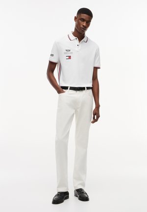 Νεαρός άνδρας όρθιος, που φοράει λευκό πόλο μπλουζάκι με λογότυπα, λευκό παντελόνι, μαύρη ζώνη και μαύρα επίσημα παπούτσια, με φόντο ένα απλό λευκό τοίχο.