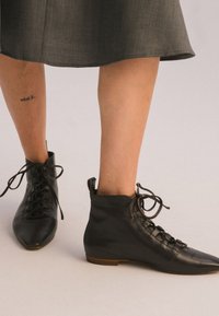 Stivaletti in pelle nera alla caviglia con punta tonda, lacci davanti e morbida texture. Suola piatta marrone e altezza alla caviglia con un dettagliato design sottile.