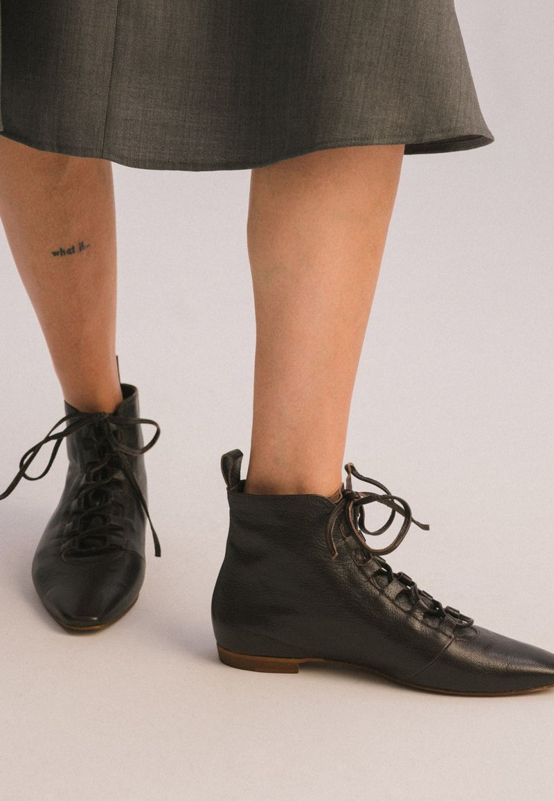 Stivaletti in pelle nera alla caviglia con punta tonda, lacci davanti e morbida texture. Suola piatta marrone e altezza alla caviglia con un dettagliato design sottile.