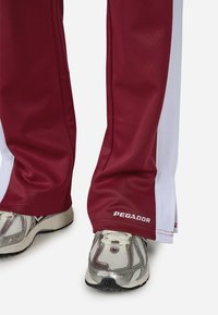 Bordowe spodnie dresowe z białymi bocznymi paskami, wykonane z gładkiego materiału. Zauważalne logo "PEGADOR" na końcu nogawki. Srebrne buty sportowe z teksturowanymi akcentami.