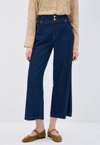 Jeans de mezclilla oscura de pierna ancha y cintura alta, con dos bolsillos frontales y acentos de botones en tono dorado en la cintura.