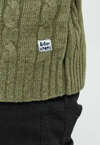 Pull en tricot vert olive avec motif en cablé et bord-côte. Présente une petite étiquette blanche avec texte bleu marine. Assorti à un pantalon noir.