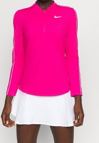 Long-sleeve pink athletic top with a quarter-zip collar and white side stripes, paired with a white flared skirt. Smooth, lightweight fabric.