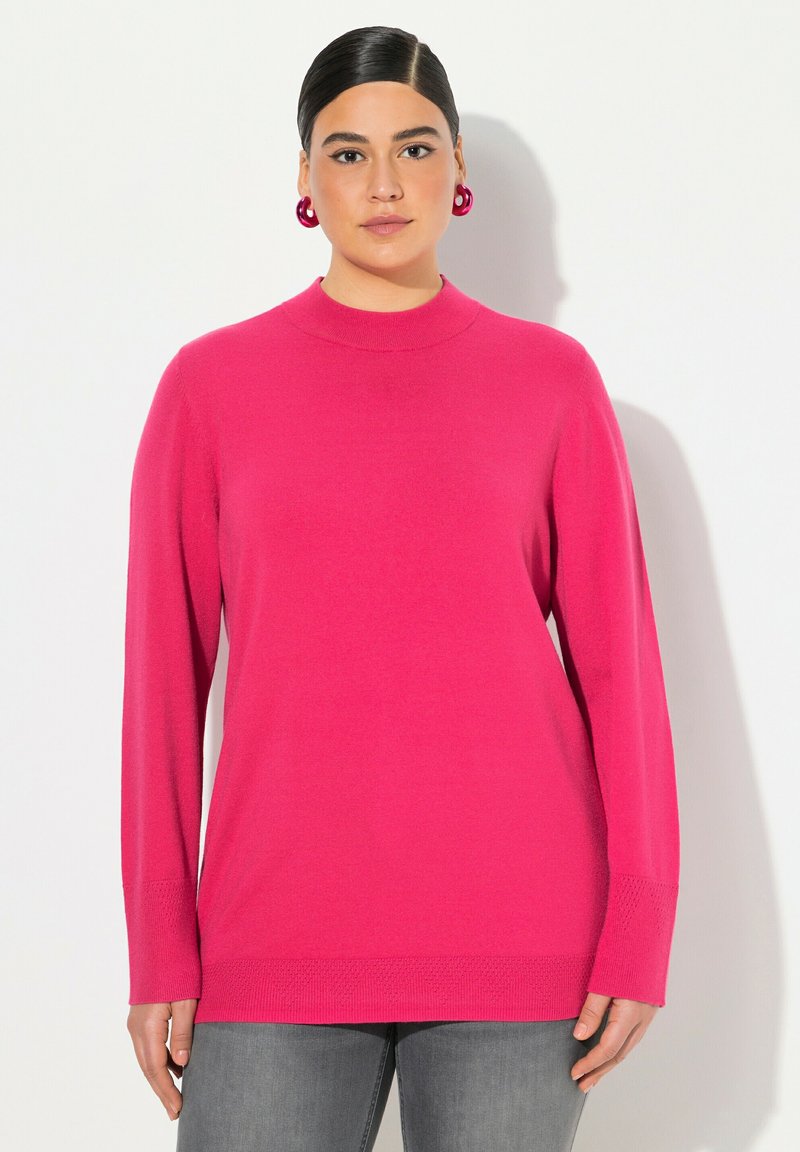 Fuchsiový svetr s vysokým límcem, dlouhými rukávy a texturovaným lemem. Nosený s šedými džíny. Hladce pletená látka; žádné viditelné vzory ani kovové prvky.
