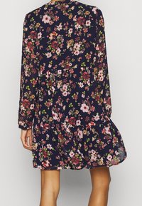 Robe fleurie en bleu marine foncé avec manches longues, jupe à volants et divers motifs floraux roses et blancs. Tissu léger, coupe décontractée.