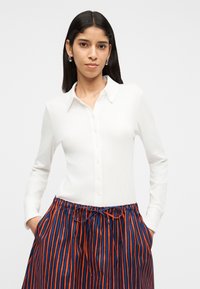 Γυναίκα με μακριά σκούρα μαλλιά που φοράει ασημένια σκουλαρίκια κρίκους, ένα λευκό πουκάμισο με κουμπιά και μακριά μανίκια, και μια φούστα με μπλε και κόκκινες ρίγες, με τα χέρια στις τσέπες.