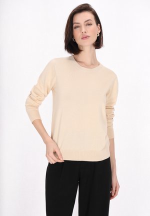 Pulover lung bej deschis cu mânecă lungă, din material moale, cu guler rotund și manșete dublate, asortat cu pantaloni negri.