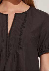 Blusa nera con scollatura a V, texture plissettata e dettagli di ricamo floreale lungo l'apertura. Maniche a palloncino e finitura in tessuto morbido.