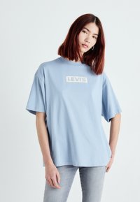 Lyseblå bomulds t-shirt med korte ærmer, rund hals, med et hvidt rektangulært Levi's-logo på forsiden. Kombineret med lysegrå bukser.